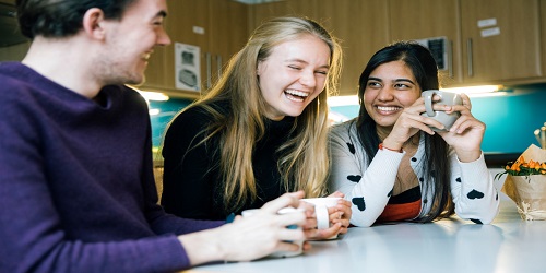 Several students of different races and genders sit around a kitchen table holding mugs and laughing.